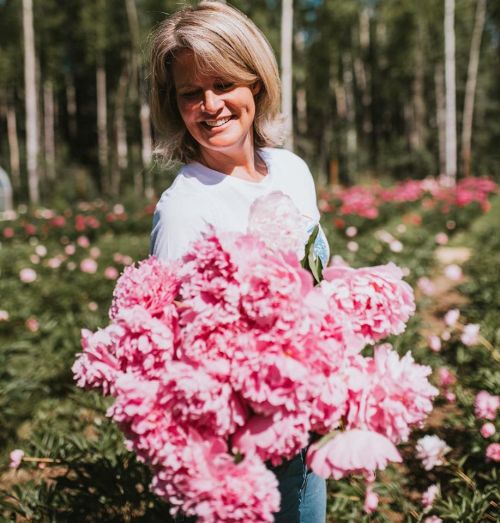 A woman smiles while holding a large bouquet of pink peonies in a flower field surrounded by greenery.