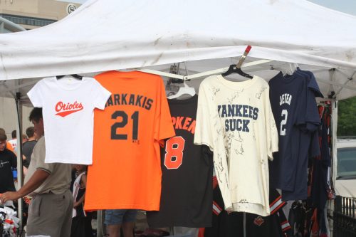 A tent displays various baseball jerseys and shirts, including Orioles, Markakis, and Yankees-themed apparel.