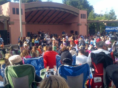 A crowd enjoying a live music performance at an outdoor stage, with people seated in lawn chairs.