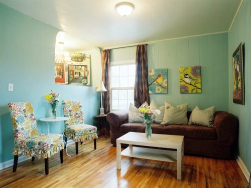 Cozy living room with a brown couch, floral chairs, colorful artwork, and a small table with a vase of flowers.