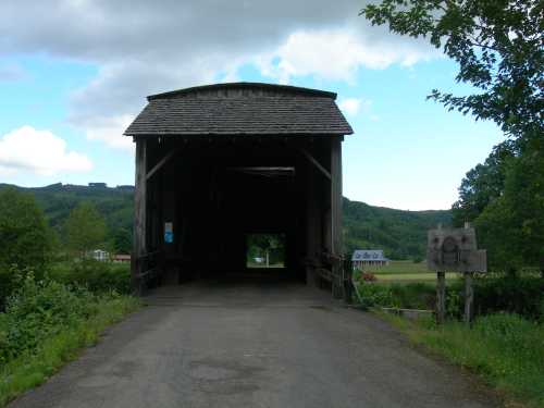 A wooden covered bridge with a gravel path leading through it, surrounded by greenery and hills in the background.