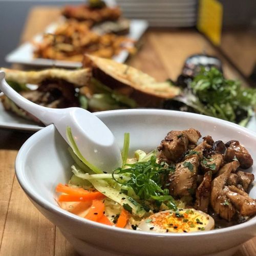 A bowl of chicken over rice with vegetables, garnished with green onions and a soft-boiled egg, on a wooden table.