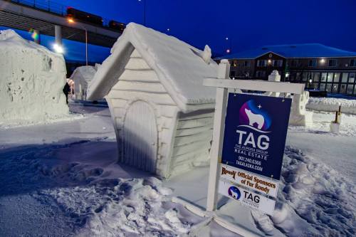 A snow sculpture of a doghouse with a sign for TAG Real Estate in a winter setting at dusk.
