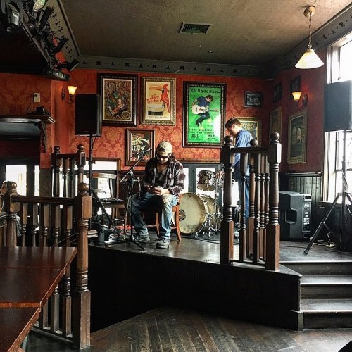 A musician sits on stage in a cozy venue, preparing for a performance with instruments and posters in the background.