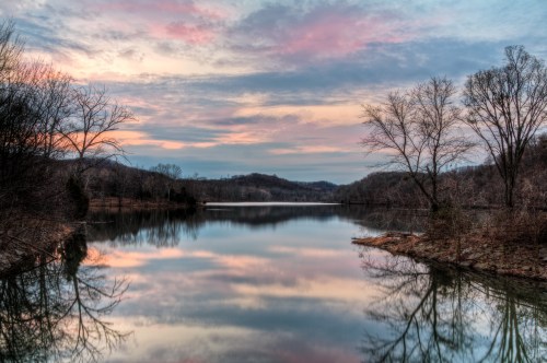 A serene lake at sunset, reflecting colorful clouds and trees along the shore, surrounded by hills.