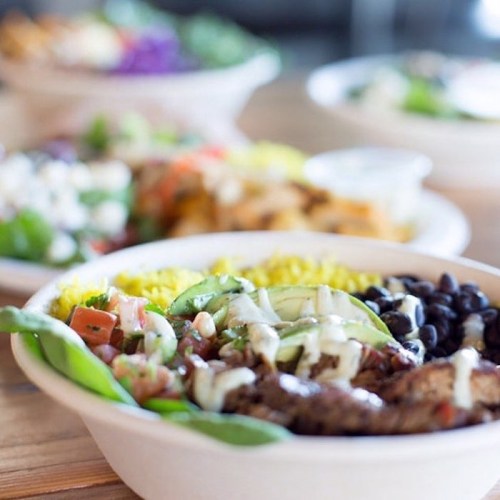 A close-up of a colorful bowl filled with rice, black beans, grilled meat, avocado, and drizzled sauce, with salads in the background.