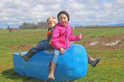 Two children sit on a large blue object in a grassy field, smiling and enjoying a sunny day outdoors.