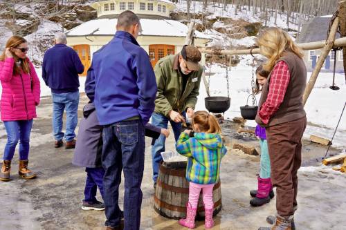A group of people gathers around a barrel, learning about maple syrup production in a snowy outdoor setting.