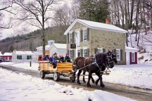 A horse-drawn wagon carries passengers through a snowy village, with historic buildings in the background.