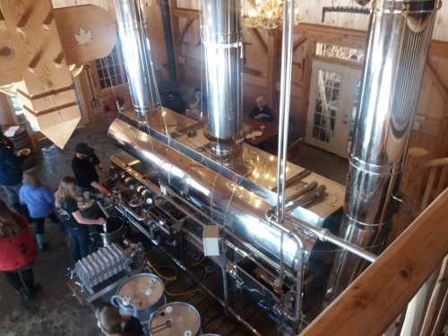 A spacious wooden barn interior with a large, shiny metal syrup evaporator and people gathered around.