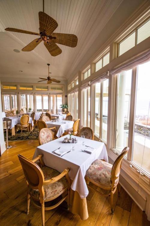 Bright dining area with large windows, wooden floors, and elegantly set tables surrounded by stylish chairs.