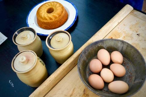 A wooden table with three jars, a round cake on a plate, and a bowl of eggs.