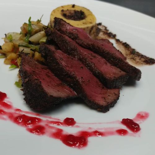 Sliced steak with a crust, accompanied by a round potato dish, vegetable medley, and a drizzle of sauce.