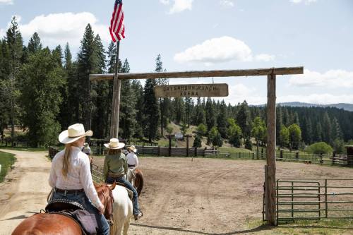Two riders on horseback approach a wooden sign for Greenhorn Ranch Arena, with trees and a flag in the background.