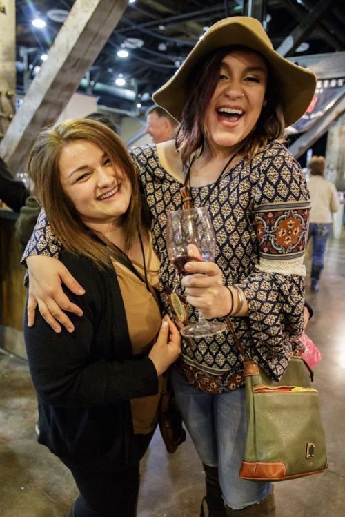 Two smiling women pose together, one holding a drink, in a lively indoor setting with rustic decor.