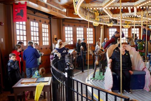 A lively carousel room with families enjoying the ride and exploring, featuring colorful decorations and wooden animals.