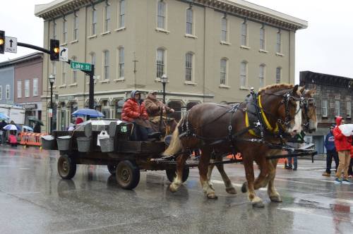 A horse-drawn cart with two people rides through a rainy street, passing by a historic building and umbrellas.