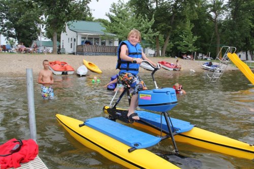 A child in a life jacket rides a yellow water bike, smiling, with people and kayaks in the background at a beach.