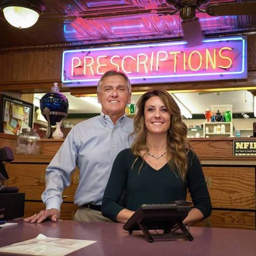 A man and woman stand behind a pharmacy counter, smiling, with a neon "PRESCRIPTIONS" sign in the background.