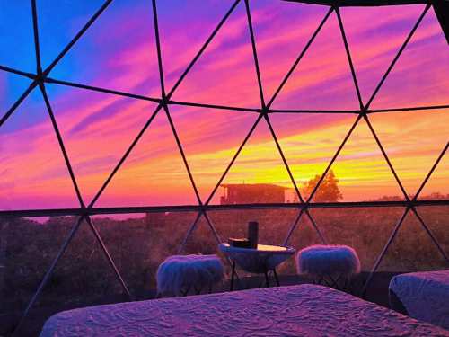 A colorful sunset viewed through a geodesic dome, with a cozy interior and soft furnishings.