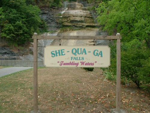 Sign for She-Qua-Ga Falls, labeled "Tumbling Waters," with a natural backdrop of trees and a waterfall.