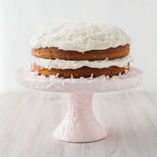 A two-layer coconut cake with white frosting, topped with shredded coconut, displayed on a pink cake stand.