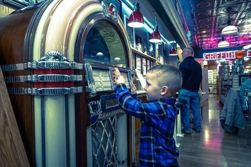 A young boy in a plaid shirt interacts with a vintage jukebox in a retro diner setting.