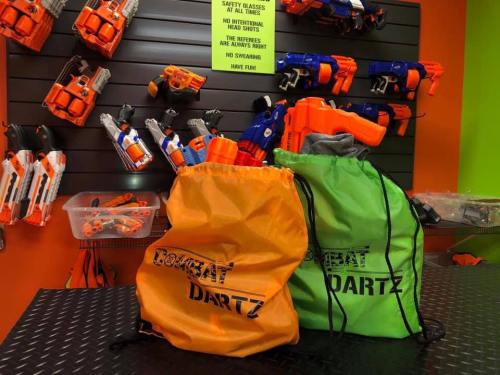 Two drawstring bags labeled "Combat Dartz" in orange and green, with toy blasters displayed on a wall behind them.