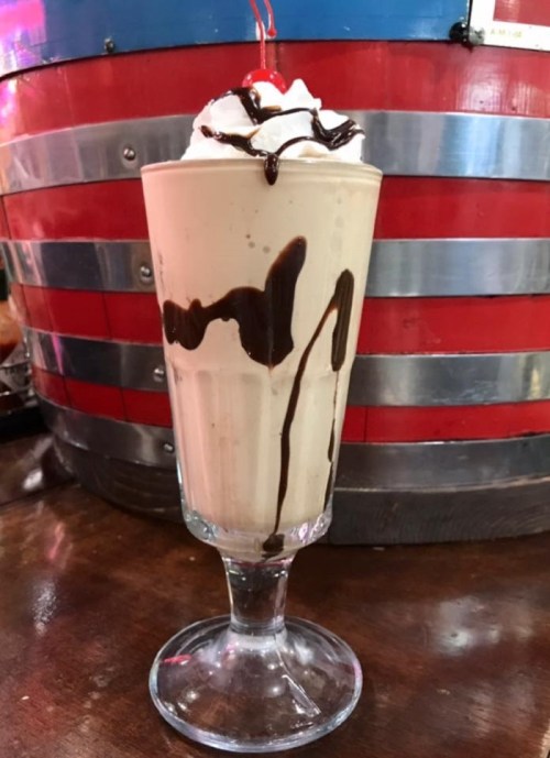 A tall glass of milkshake topped with whipped cream, a cherry, and drizzled with chocolate sauce.