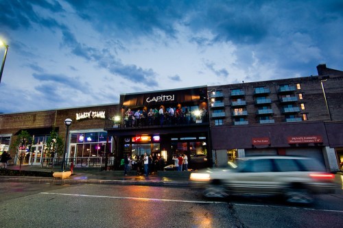 A lively street scene at dusk featuring a bar with a balcony and nearby buildings, with a car passing by.