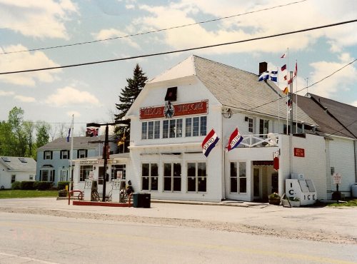 A white building with flags and signs, featuring a large front window and a small gas station in front.