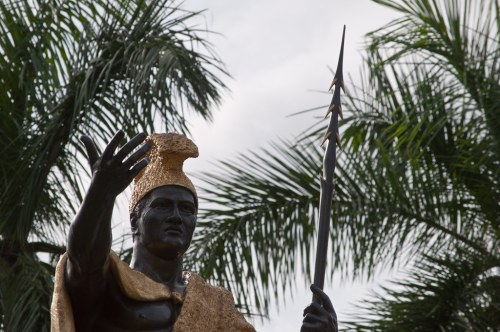 Statue of a warrior in a golden headdress, holding a spear, surrounded by palm trees.