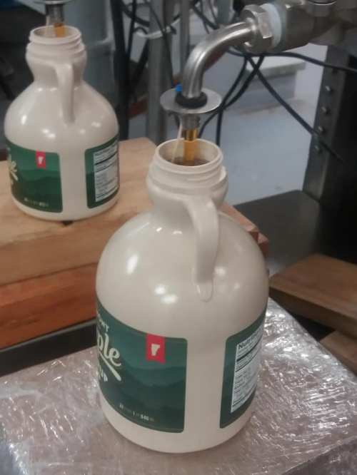 A close-up of a maple syrup jug being filled from a spout in a production facility.