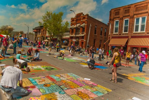 A vibrant street festival with artists creating colorful chalk art on pavement, surrounded by spectators and historic buildings.