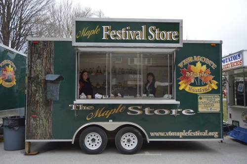 A green food truck with "Maple Festival Store" signage, featuring two smiling vendors inside.
