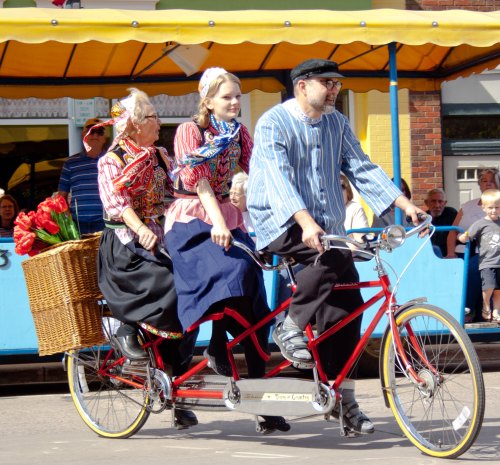 Three people in traditional attire ride a tandem bicycle, with a basket of flowers, during a festive event.
