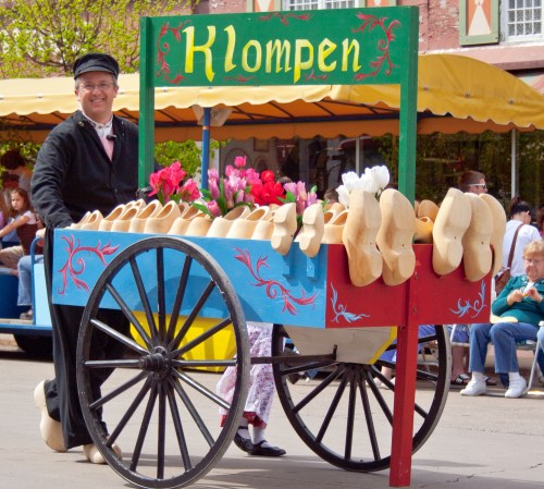A man in traditional attire stands beside a colorful cart filled with wooden clogs and flowers at a festive event.