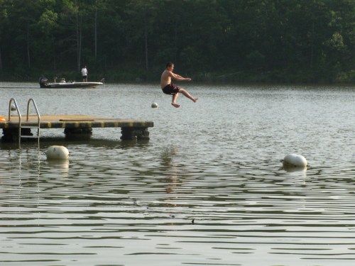 A person jumps off a dock into a lake, with a boat in the background and calm water rippling around them.