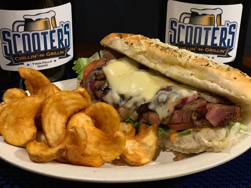 A hearty sandwich with meat, cheese, and veggies, served with crispy potato chips and two bottles of Scooters drink.