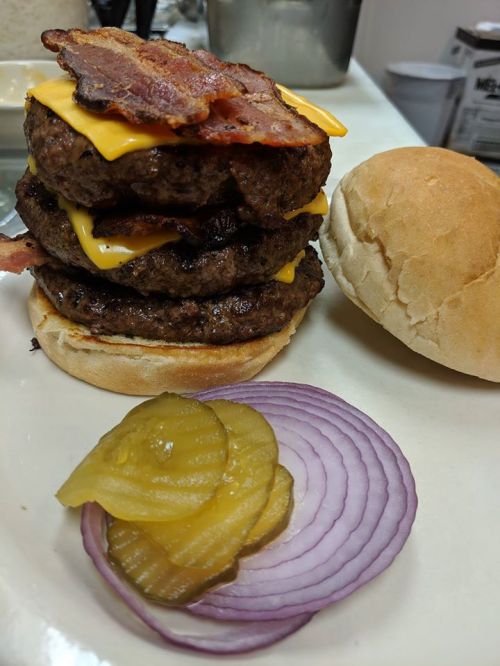 A towering burger with three beef patties, melted cheese, bacon, and a side of pickles and red onion slices.