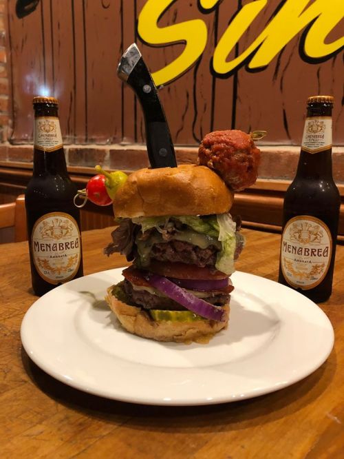 A towering burger with a knife on top, surrounded by two bottles of Menabrea beer on a wooden table.