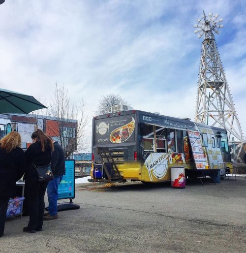 A food truck with a menu displayed, parked near a windmill, with customers ordering food.