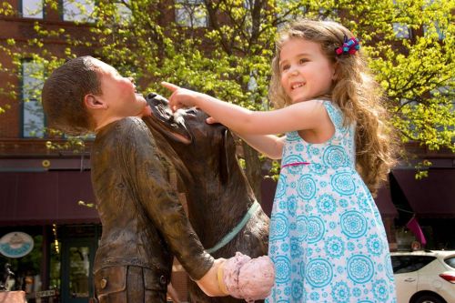 A girl in a blue dress playfully points at a bronze statue of a boy and a dog while holding ice cream.