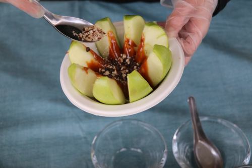 A person in a glove adds toppings to a bowl of sliced green apples drizzled with caramel and sprinkled with nuts.