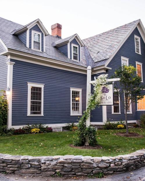 A blue farmhouse with a sign reading "Solo Farm & Table," surrounded by greenery and flowers.