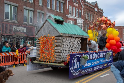 A parade float resembling a log cabin, decorated with autumn leaves and balloons, with people preparing it for the event.