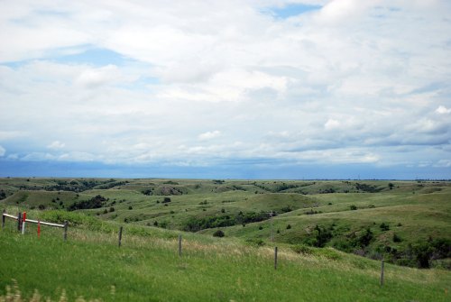 Lush green hills under a cloudy sky, with a fence line in the foreground and rolling landscape in the background.