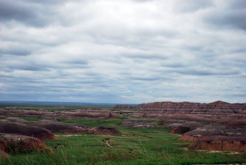 A vast landscape of rolling hills and grassy plains under a cloudy sky, with colorful rock formations in the distance.