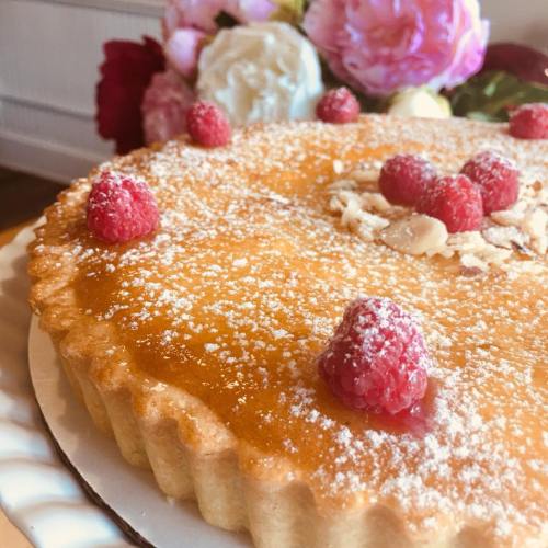A golden tart topped with raspberries and almond slices, dusted with powdered sugar, surrounded by pink flowers.