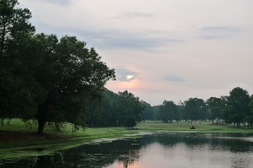 A serene landscape featuring a calm pond, trees, and a soft sunrise peeking through the clouds.